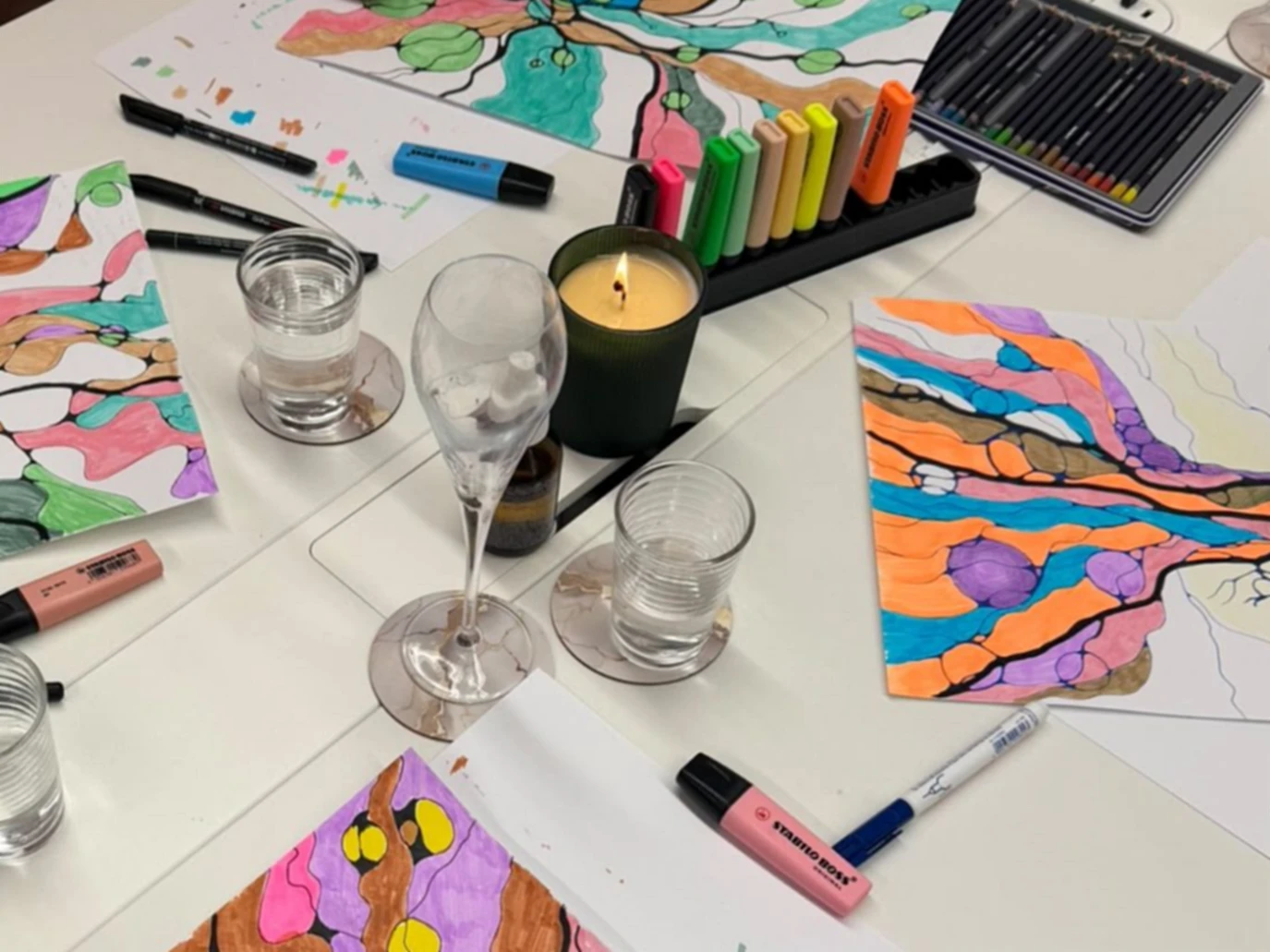 A white table seen from above, with colourful tree drawings in progress, markers, a lit candle and glasses of water